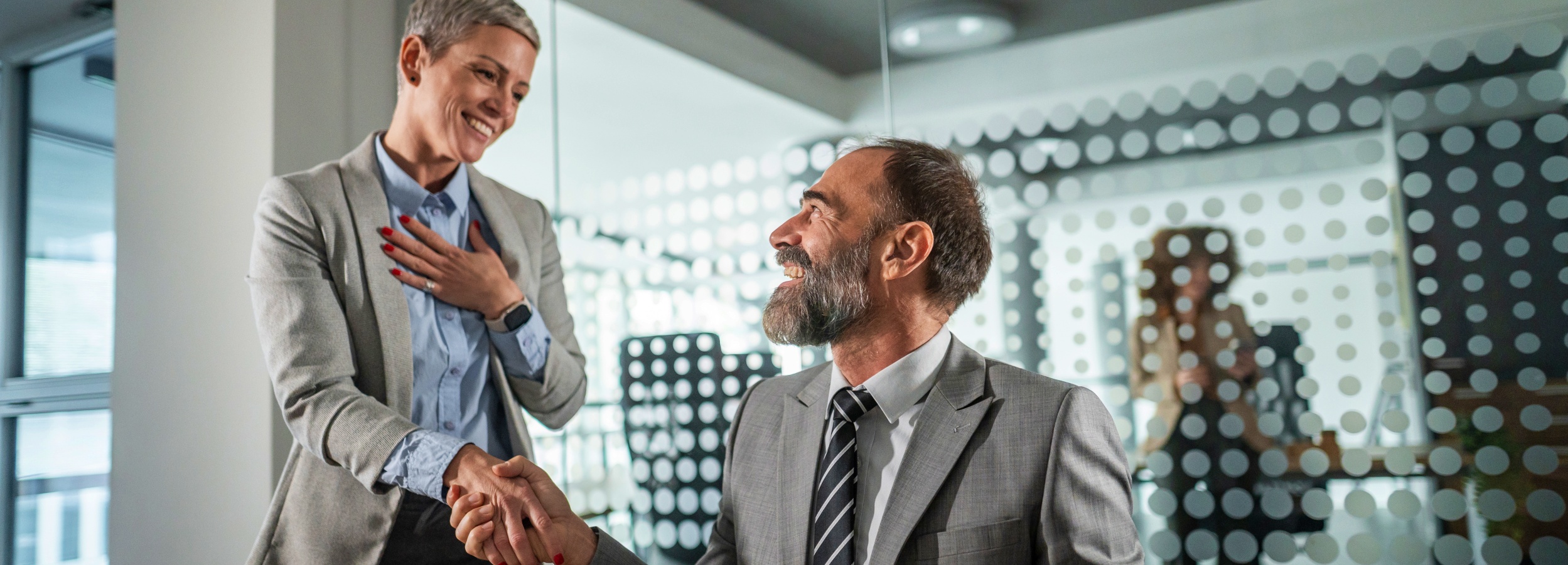 Das Bild zeigt eine geschäftliche Interaktion zwischen einer Frau und einem Mann in einem modernen Büro.

Das Bild zeigt zwei Personen: eine Frau mit kurzem, grauem Haar in einem grauen Blazer und einer hellblauen Bluse, die einem Mann die Hand gibt. Der Mann, der ebenfalls einen grauen Anzug mit gestreifter Krawatte trägt, hat einen grauen Bart und lächelt. Die Frau hält ihre linke Hand auf ihrer Brust, was auf Dankbarkeit oder Wertschätzung hindeutet. Im Hintergrund befindet sich eine Glaswand mit einem Punktmuster, durch die man verschwommen eine weitere Person an einem Schreibtisch erkennen kann. Die Atmosphäre wirkt professionell und freundlich, mit einer positiven und hoffnungsvollen Stimmung, die durch das Lächeln der beiden Hauptpersonen unterstrichen wird. Das Licht ist hell und gleichmäßig, was die moderne und saubere Ästhetik des Büros betont.