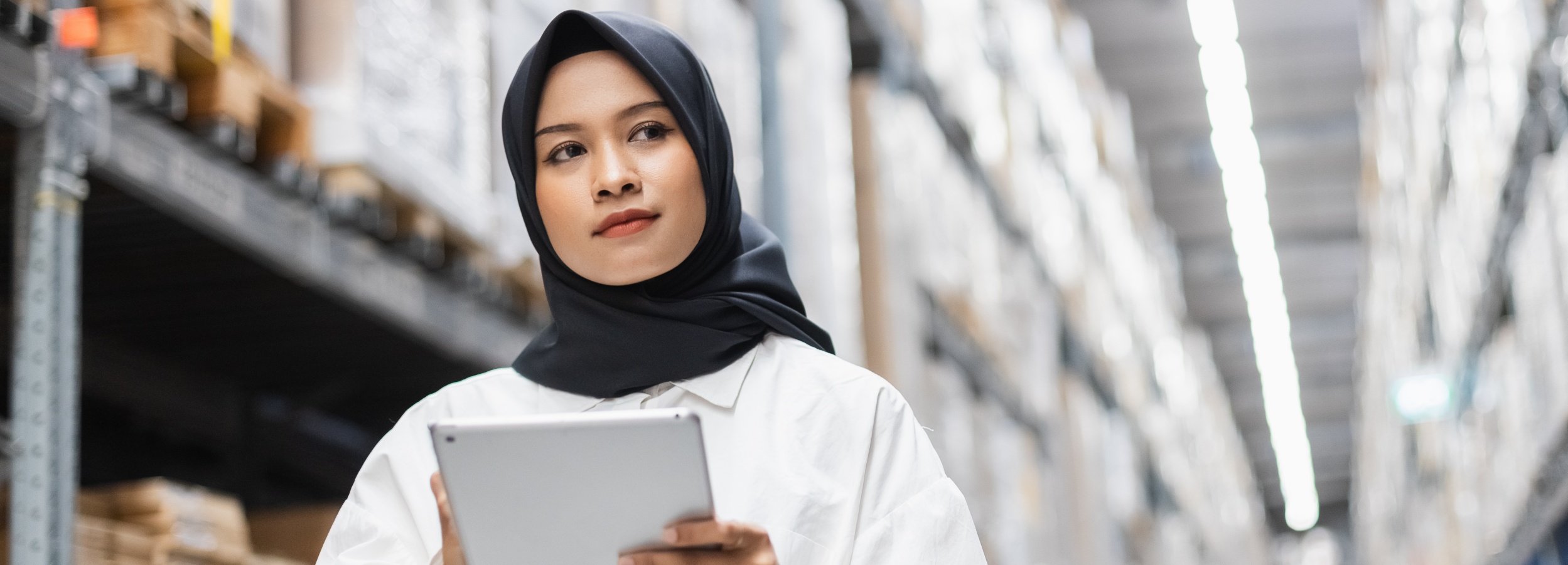 Das Bild zeigt eine junge Frau in einem Lager, die ein Tablet in der Hand hält.

Das Bild ist ein Panoramabild, das eine Nahaufnahme einer jungen Frau zeigt, die eine schwarze Hijab und ein weißes Hemd trägt. Sie blickt nach rechts und hält ein silbernes Tablet in ihren Händen. Hinter ihr erstreckt sich ein breiter Gang eines Lagers, flankiert von hohen Regalen, die mit zahlreichen Kartons gefüllt sind. Die Regale sind in einer symmetrischen Anordnung angeordnet und erzeugen eine tiefe Perspektive. Über den Regalen sind helle Leuchtstoffröhren angebracht, die den Raum gleichmäßig ausleuchten und für eine klare, fast klinische Atmosphäre sorgen. Die Beleuchtung ist hell und diffus, was auf eine professionelle und organisierte Umgebung hindeutet. Die Stimmung des Bildes ist ruhig und konzentriert. Die Frau scheint in ihre Aufgabe vertieft zu sein, was sie zu einer aktiven und kompetenten Person in dieser Arbeitsumgebung macht. Die weiche Schärfe im Hintergrund lenkt den Fokus auf die Frau und ihre Tätigkeit, während die gestaffelten Regale eine klare räumliche Tiefe vermitteln.
