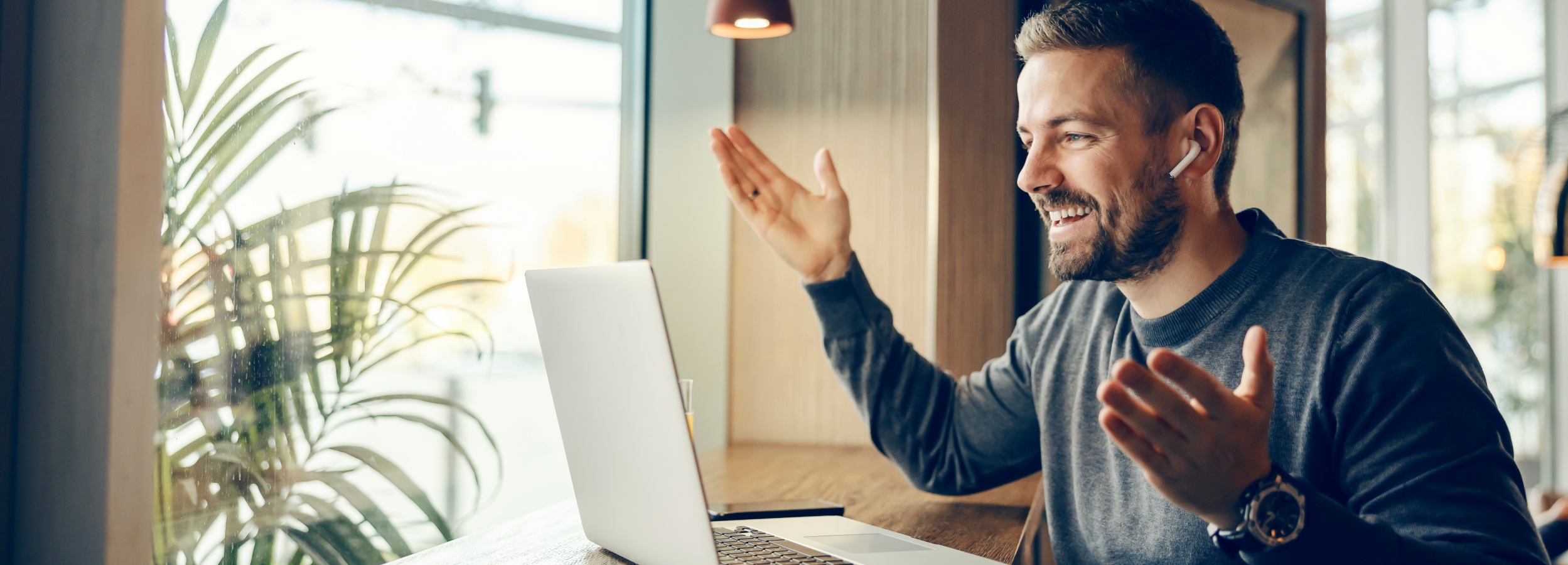Junger Mann im Internetkaffee begrüßt freudig teilnehmer einer Videokonferenz. Seine Hände sind erhoben und er hat Kopfhörer auf.