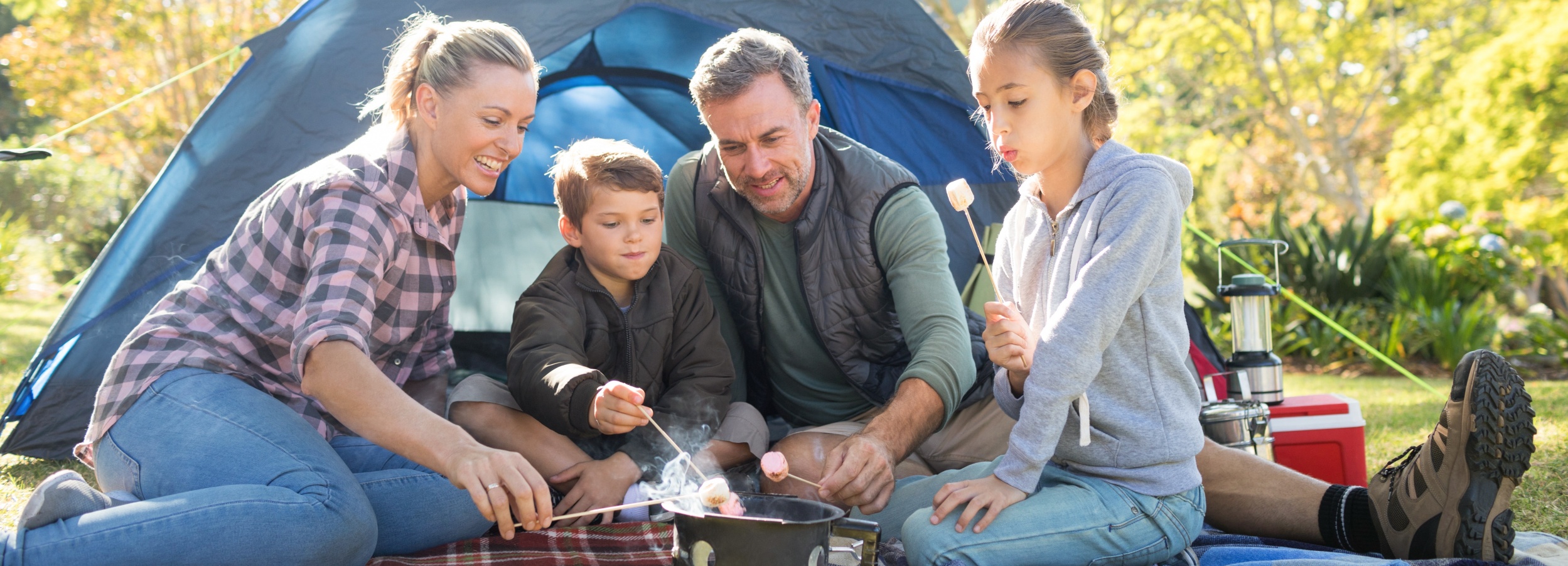 Vierköpfige Familie sitzt im Urlaub vor ihrem Zelt im Wald und grillt Marschmallows.