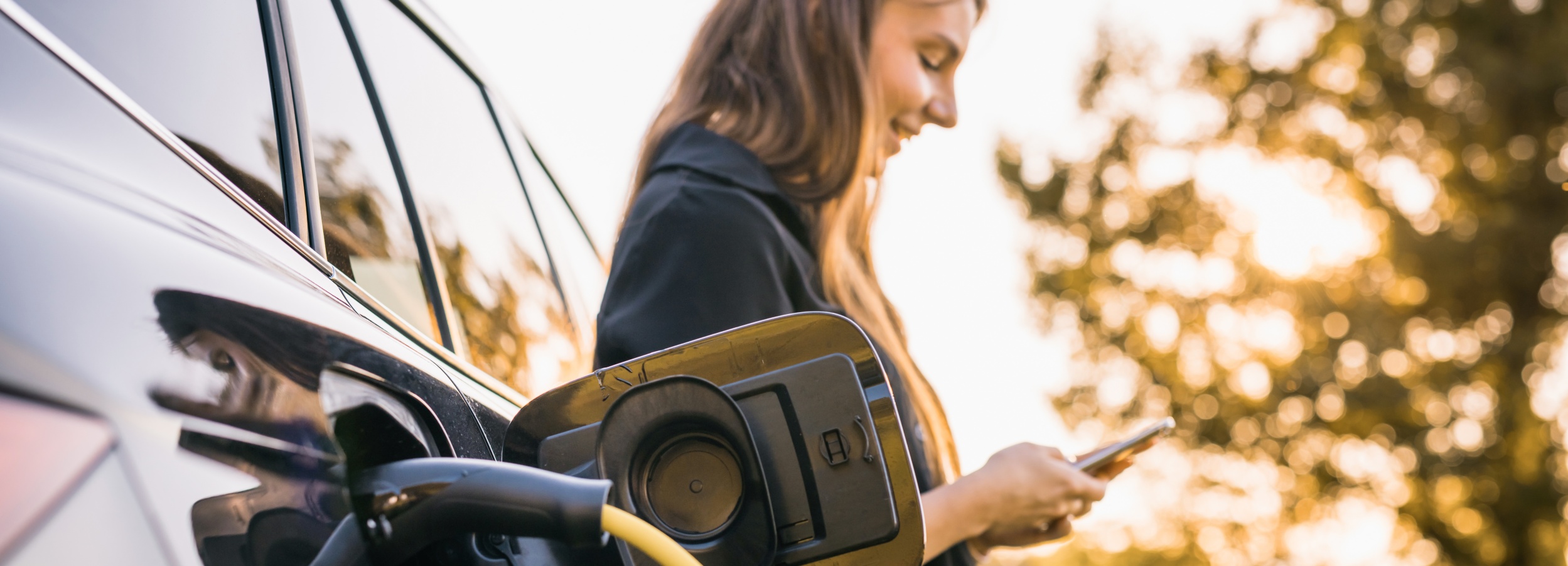 Junge Frau lädt ihr e-Auto und blickt dabei auf ihre Smartphone-App.