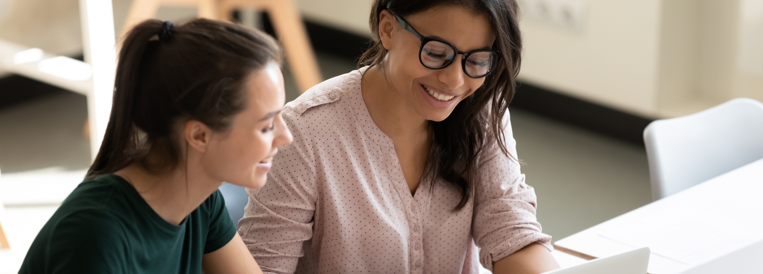Das Bild zeigt zwei lächelnde Frauen, die gemeinsam an einem Laptop arbeiten und eine positive und kollaborative Arbeitsatmosphäre ausstrahlen.

Im Detail sieht man zwei Frauen, die eng nebeneinander an einem weißen Tisch sitzen. Die Frau links trägt ein dunkelgrünes T-Shirt und hat ihre braunen Haare zu einem Pferdeschwanz gebunden. Sie lacht leicht und schaut auf den Laptop, den die andere Frau hält. Die Frau rechts trägt eine hellrosa Bluse mit kleinen weißen Punkten und eine Brille. Sie lächelt breit und scheint konzentriert auf den Laptop zu sein. Ihr dunkles Haar fällt ihr über die Schultern. Der Laptop ist im Bildausschnitt nur teilweise sichtbar. Im Hintergrund sieht man unscharf die Beine von Stühlen und einen hellen Raum. Die allgemeine Atmosphäre ist freundlich, positiv und konzentriert. Die Stimmung wird durch die lächelnden Gesichter der Frauen und die Nähe zueinander vermittelt. Das Bild strahlt Zusammenarbeit und positive Arbeitsbeziehungen aus.