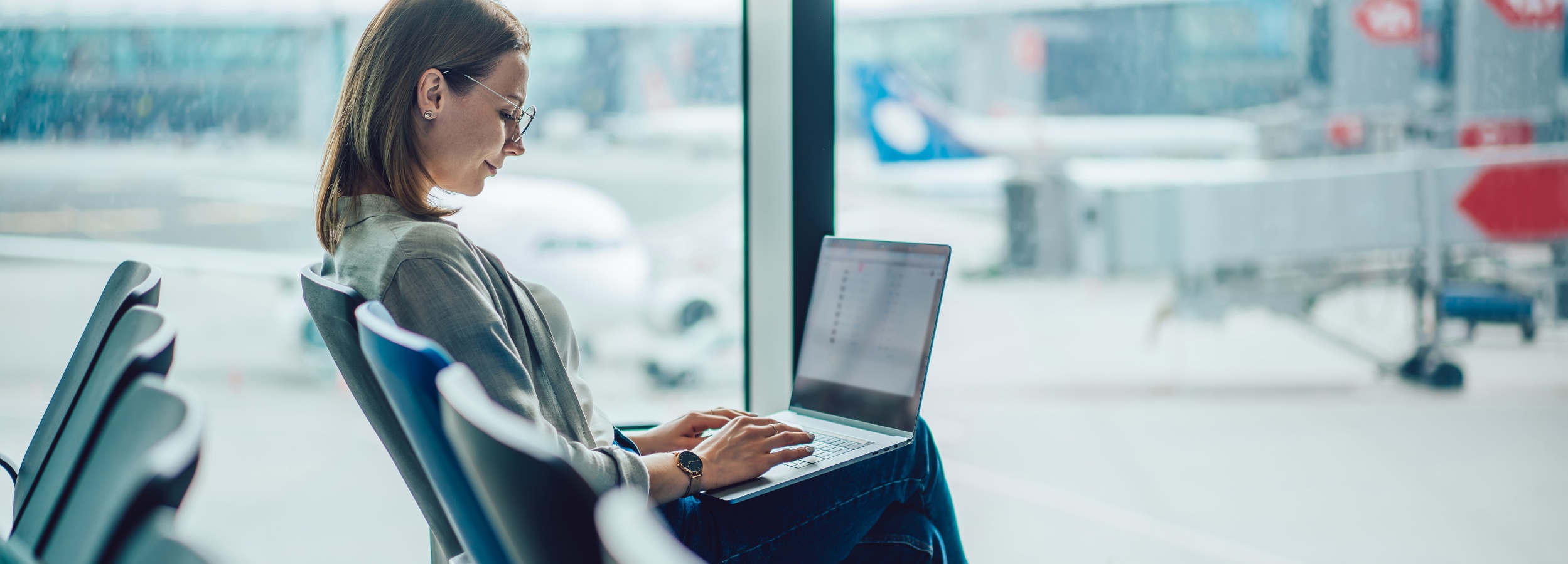 Das Bild zeigt eine Frau, die konzentriert an ihrem Laptop in einem Flughafen-Wartezimmer arbeitet.

Im Bild ist eine junge Frau mit braunen, schulterlangen Haaren zu sehen, die eine graue Bluse und Jeans trägt. Sie trägt eine Brille und eine Uhr am Handgelenk. Sie sitzt auf einem bequemen Stuhl in einem Flughafen-Wartezimmer und arbeitet an ihrem Laptop, der auf ihrem Schoß liegt. Ihre Hände tippen konzentriert auf die Tastatur. Ihr Blick ist auf den Bildschirm gerichtet und ihr Gesichtsausdruck wirkt fokussiert und konzentriert. Im Hintergrund ist durch ein großes Fenster die Rollbahn eines Flughafens mit Flugzeugen und Gepäckwagen unscharf zu sehen. Die Atmosphäre ist ruhig und konzentriert, obwohl der Ort ein geschäftiger Flughafen ist. Die Farben sind gedämpft, die Lichtverhältnisse hell und natürlich. Der gesamte Eindruck ist einer von konzentrierter Arbeit in einer öffentlichen, aber ruhigen Umgebung.