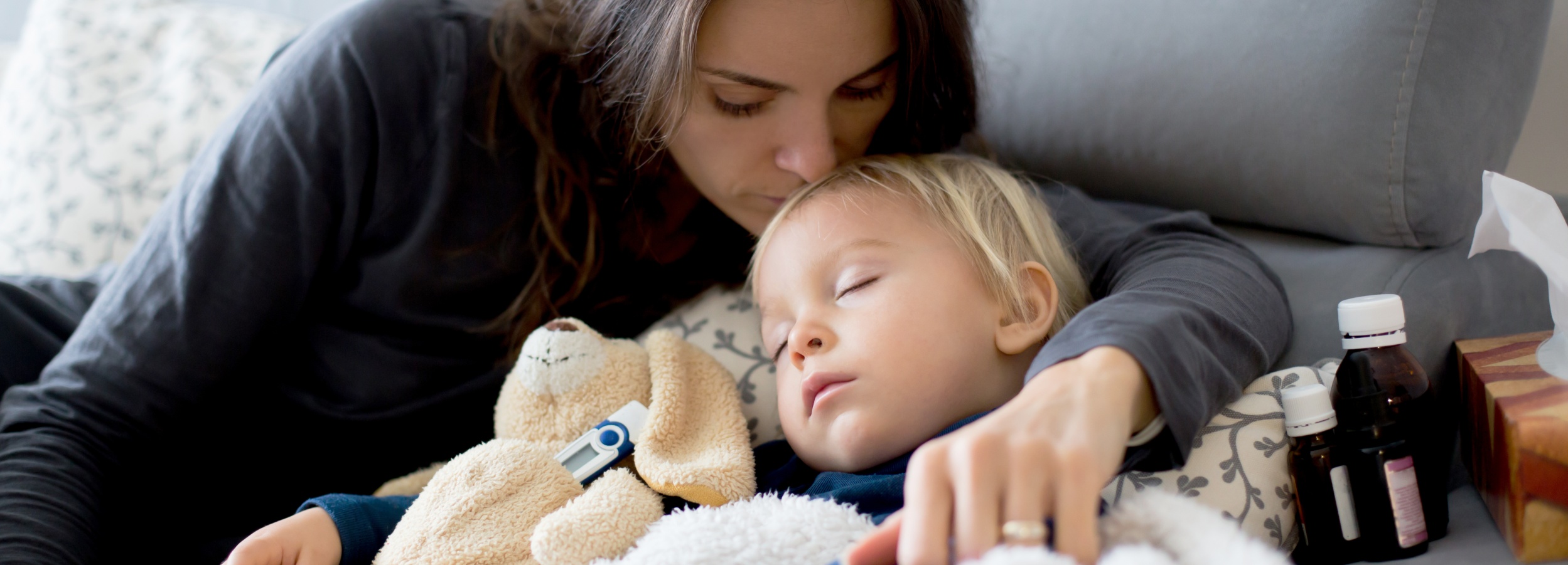 Das Bild zeigt eine Mutter, die ihr krankes Kleinkind liebevoll auf der Couch tröstet.

Das Bild zeigt eine junge Frau mit langen, braunen Haaren, die ein kleines Kind mit blonden Haaren auf der Couch im Arm hält. Das Kind schläft und liegt eingekuschelt in eine Decke. Neben dem Kind liegt ein beigefarbenes Stofftier in Form eines Hundes, an dem ein digitales Fieberthermometer befestigt ist. Die Mutter beugt sich liebevoll über das Kind und legt ihr einen Kuss auf die Stirn. Ihr Gesichtsausdruck ist sanft und besorgt. Ihre Hand ruht tröstend auf dem Kind. Im Hintergrund sieht man einen Teil eines grauen Sofas und neben dem Kind stehen mehrere kleine Flaschen, die vermutlich Medikamente enthalten, sowie ein Paket Taschentücher. Die Atmosphäre des Bildes ist warm, ruhig und emotional, geprägt von Fürsorge und Zärtlichkeit. Die Farben sind gedämpft und natürlich. Das Gesamtbild vermittelt eine Stimmung von Geborgenheit und Fürsorge in einer Situation von Krankheit und Sorge um das Kind.