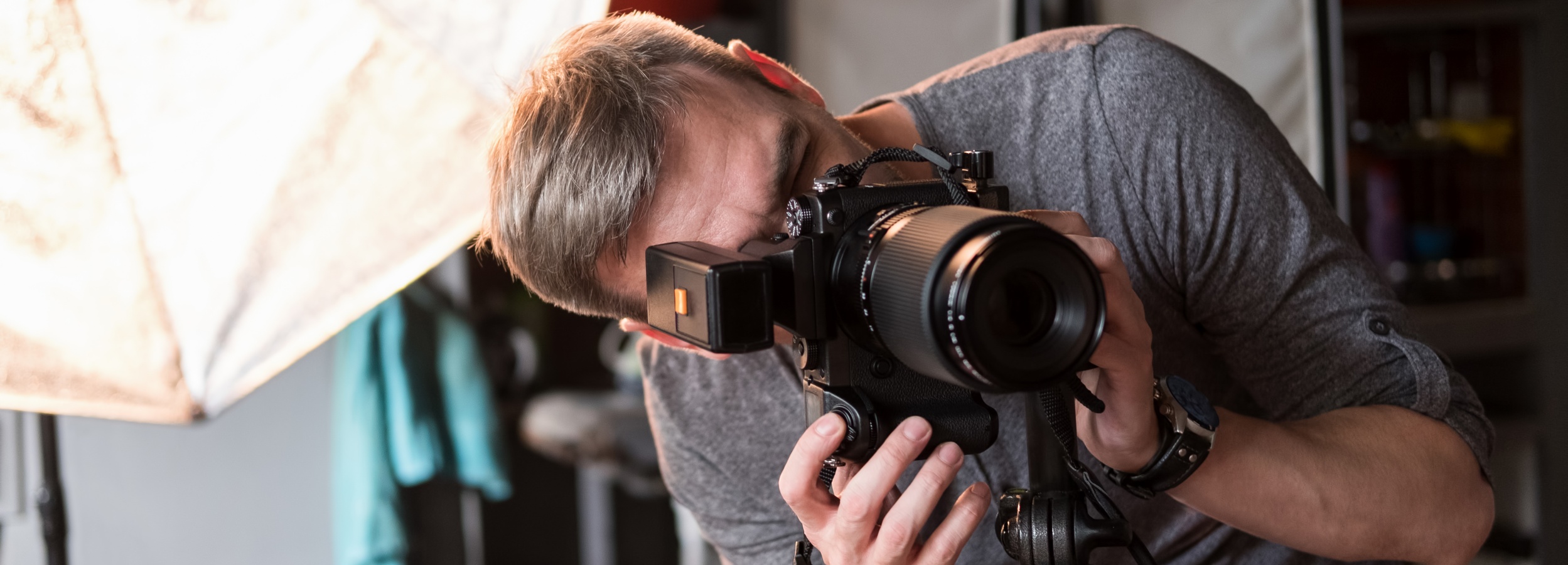 Das Bild zeigt einen konzentrierten Fotografen, der durch eine professionelle Kamera hindurch in die Linse blickt.

Im Detail zeigt das Bild einen Mann mittleren Alters mit kurzen, grau melierten Haaren. Er trägt ein schlichtes, graues T-Shirt und eine Armbanduhr mit Lederarmband. Sein Gesichtsausdruck ist konzentriert und ernst; er ist ganz auf das Fotografieren fokussiert. Er hält eine große, professionelle Spiegelreflexkamera mit einem langen Teleobjektiv. An der Kamera ist ein externes Blitzgerät angebracht. Die Kamera steht auf einem Stativ. Im Hintergrund ist unscharf ein Fotostudio mit Beleuchtungsequipment (ein großer, weicher Diffusor ist teilweise sichtbar) und weitere, nicht genau erkennbare Gegenstände zu sehen. Die Atmosphäre ist ruhig und konzentriert, fast etwas düster, aufgrund der gedämpften Beleuchtung. Die Stimmung ist professionell und intensiv, die Konzentration des Fotografen steht im Vordergrund.