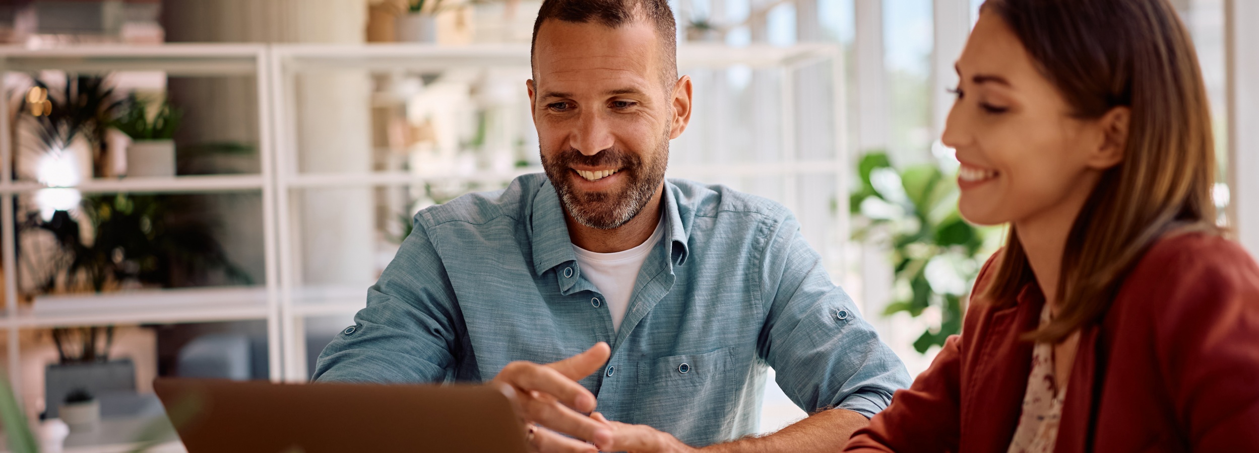 Das Bild zeigt einen Mann und eine Frau, die an einem Tisch vor einem Laptop sitzen und sich angeregt unterhalten.

Im Vordergrund sind ein Mann und eine Frau zu sehen, die an einem Tisch sitzen. Der Mann, zu seiner Rechten, hat einen leichten Bart und trägt ein hellblaues Hemd über einem weißen T-Shirt. Er lächelt und gestikuliert mit seinen Händen, was darauf hindeutet, dass er etwas erklärt oder betont. Die Frau, zu seiner Linken, hat braune Haare und trägt eine rostrote Jacke über einem gemusterten Oberteil. Sie lächelt ebenfalls und scheint aufmerksam zuzuhören.

Zwischen den beiden Personen befindet sich ein Laptop, der auf dem Tisch steht. Vor dem Laptop liegt ein Blatt Papier. Der Tisch selbst scheint aus Holz zu sein.

Im Hintergrund ist ein Raum zu sehen, der hell und modern wirkt. Ein Regal mit Pflanzen und dekorativen Elementen ist erkennbar. Durch das Fenster im Hintergrund sind verschwommene Umrisse von Gebäuden und Fahrzeugen zu erkennen, was darauf hindeutet, dass sich die Szene in einem urbanen Umfeld abspielt.

Die Atmosphäre des Bildes ist positiv und professionell. Die Stimmung ist freundlich und aufmerksam. Die Interaktion zwischen den beiden Personen deutet auf ein produktives Gespräch oder eine Zusammenarbeit hin. Das helle und moderne Umfeld verstärkt den Eindruck einer positiven und dynamischen Arbeitsatmosphäre.