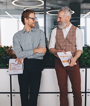 Zwei Geschäftsmänner, einer jünger, der andere älter mit einem grauen Bart, unterhalten sich locken im Flur eines Bürogebäudes.